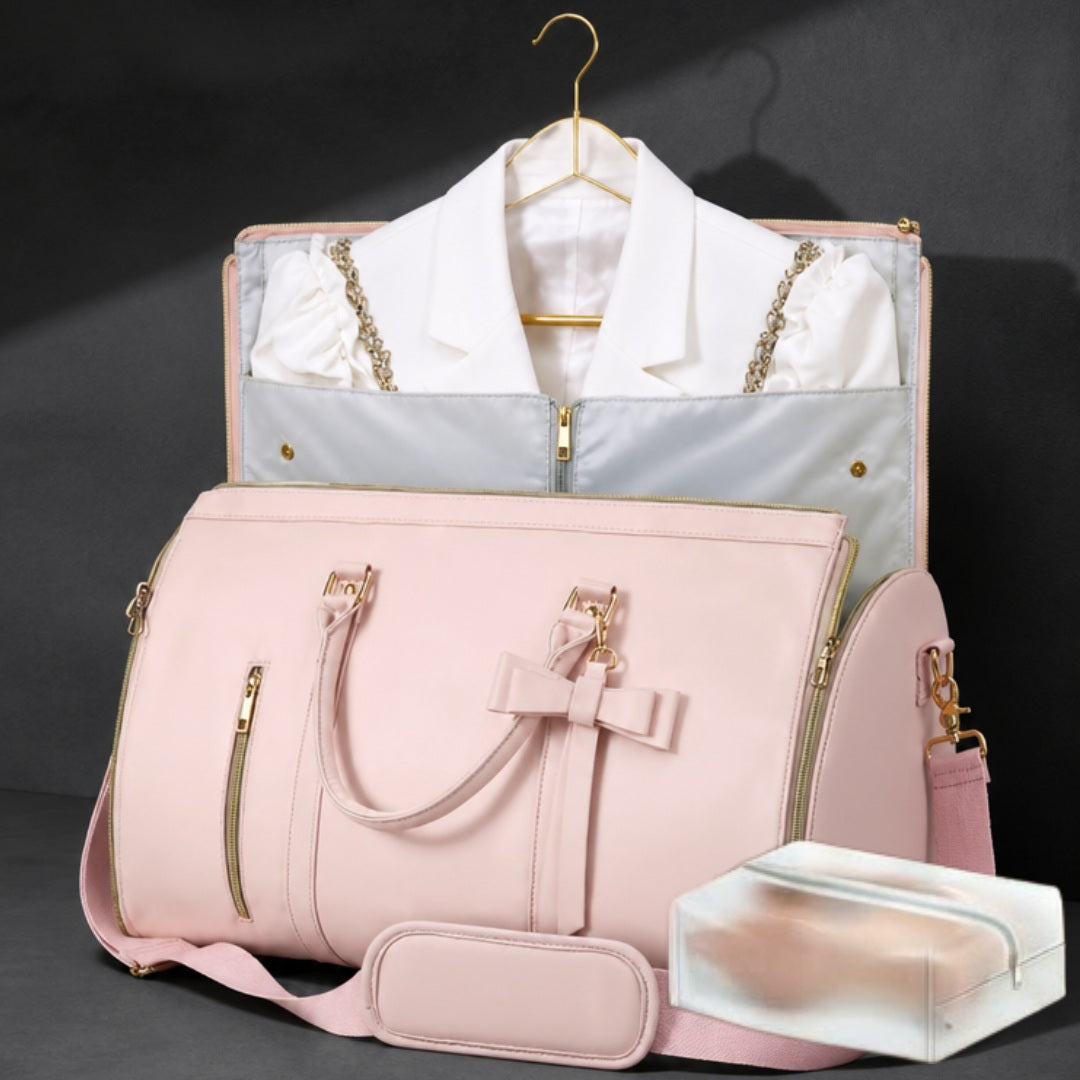 A pink structured duffel bag with gold-tone hardware and a shoulder strap, shown in front of a matching garment bag containing a white collared shirt. A small clear plastic case sits in the lower right on a dark studio background.