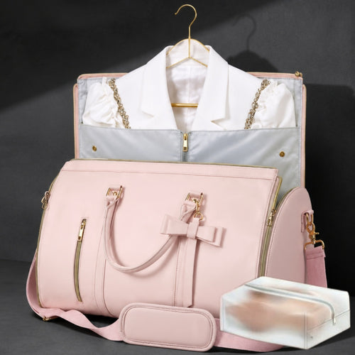 A pink structured duffel bag with gold-tone hardware and a shoulder strap, shown in front of a matching garment bag containing a white collared shirt. A small clear plastic case sits in the lower right on a dark studio background.