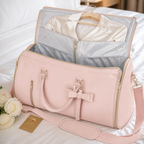 A light pink travel bag with gold zippers and a bow detail, shown open on a white bed with a white shirt inside. A small gold card and white roses are placed beside it.